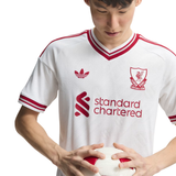 A white Adidas Liverpool FC home soccer jersey with red trim, featuring the club crest and Standard Chartered sponsor logo, worn by a model holding a soccer ball.