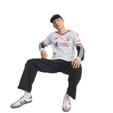 A man wearing a white Adidas soccer jersey with red and black trim, black sweatpants, white sneakers, and a black cap, posed against a solid white background.