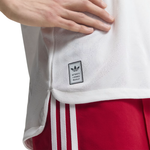 Close-up of a white Adidas athletic jersey with a small black brand patch near the hem, worn with red shorts featuring white side stripes. A hand is visible holding the jersey.