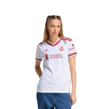 A woman modeling a white Adidas Liverpool football jersey with red trim, featuring the club's crest and sponsor logos, paired with blue jeans against a plain white background.