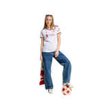 A woman modeling a white Adidas soccer jersey with red trim and sponsor logos, paired with blue jeans, white sneakers, and a red-and-white scarf, posed against a plain white background with a soccer ball at her foot.