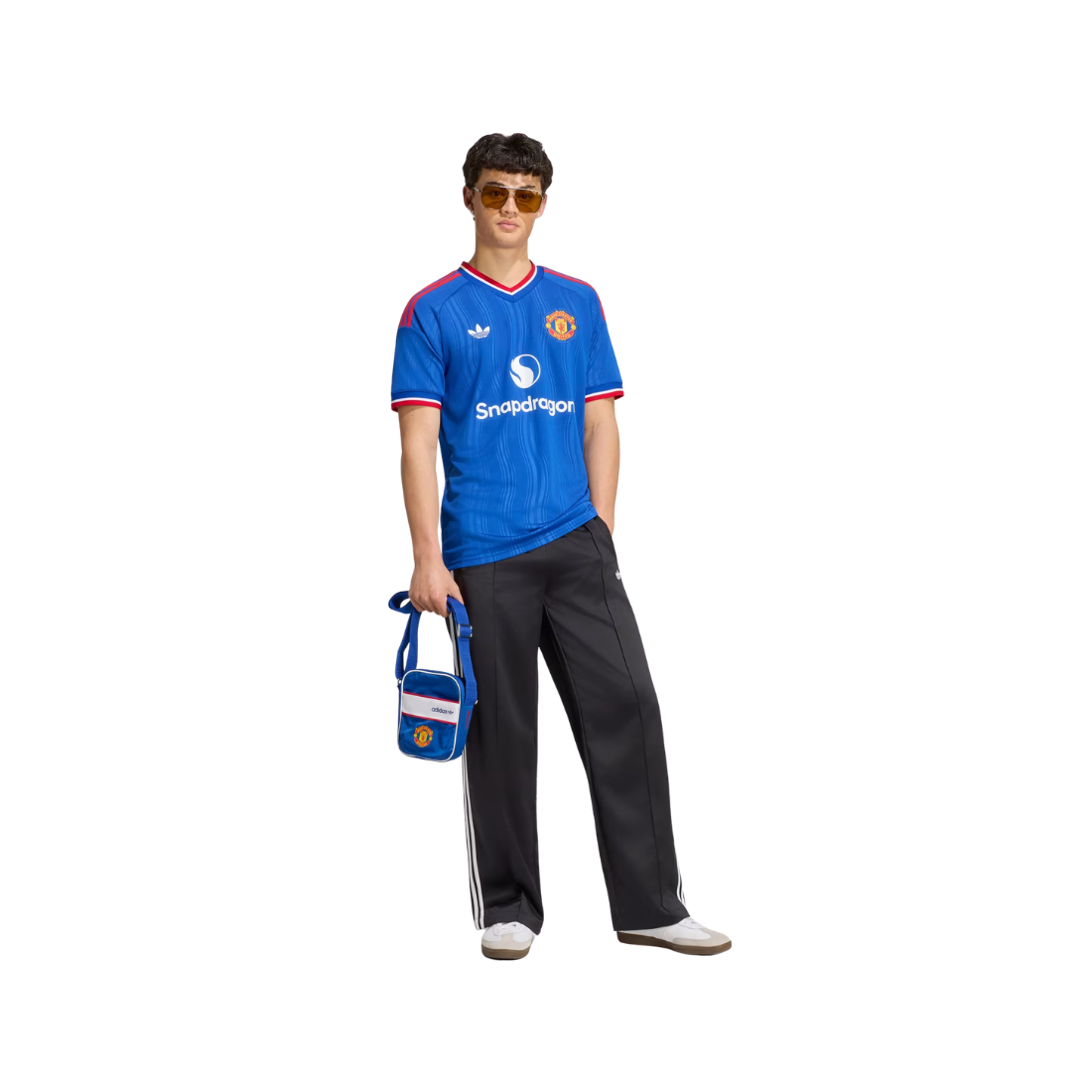 A man modeling a blue Manchester United soccer jersey with red and white trim, black track pants, sunglasses, and holding a small blue and white bag against a plain white background.