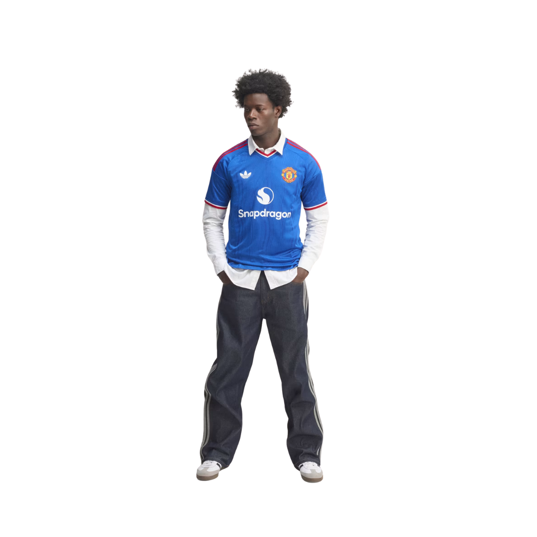 A full-body model wearing a blue Manchester United football jersey with 'Snapdragon' sponsor logo, layered over a white long-sleeve shirt, paired with dark gray track pants and white sneakers, set against a plain white background.