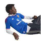 A man wearing a blue Manchester United soccer jersey with white long-sleeve undershirts and dark shorts, posed dynamically against a plain white background.