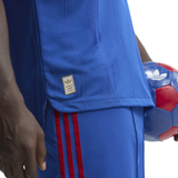 Close-up of a person wearing a royal blue Adidas soccer jersey and matching shorts with red stripes, holding a red and blue soccer ball against a white background.