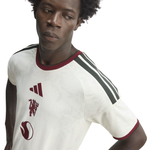 A close-up of a man wearing an Adidas Manchester United home soccer jersey. The jersey is off-white with dark red trim on the crew neck and sleeve cuffs, featuring three dark green stripes on the shoulders and the club's crest on the left chest.