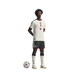 A full-body model wearing a white Adidas soccer jersey with dark green and red trim, matching dark green shorts, white socks, and white soccer cleats, standing next to a soccer ball on a plain white background.