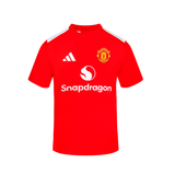 A red short-sleeve soccer jersey Junior with white shoulder stripes, featuring the Manchester United crest on the left chest, the white Adidas logo on the right chest, and the white Snapdragon sponsor logo across the center.